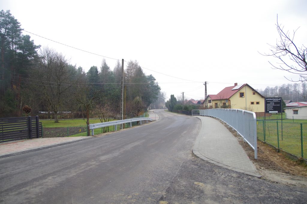 Widok przebudowanego odcinka drogi od strony skrzyżowania do centrum wsi. Po prawej stronie za chodnikiem szara metalowa barierka, a po lewej drogowa barierka ochronna.