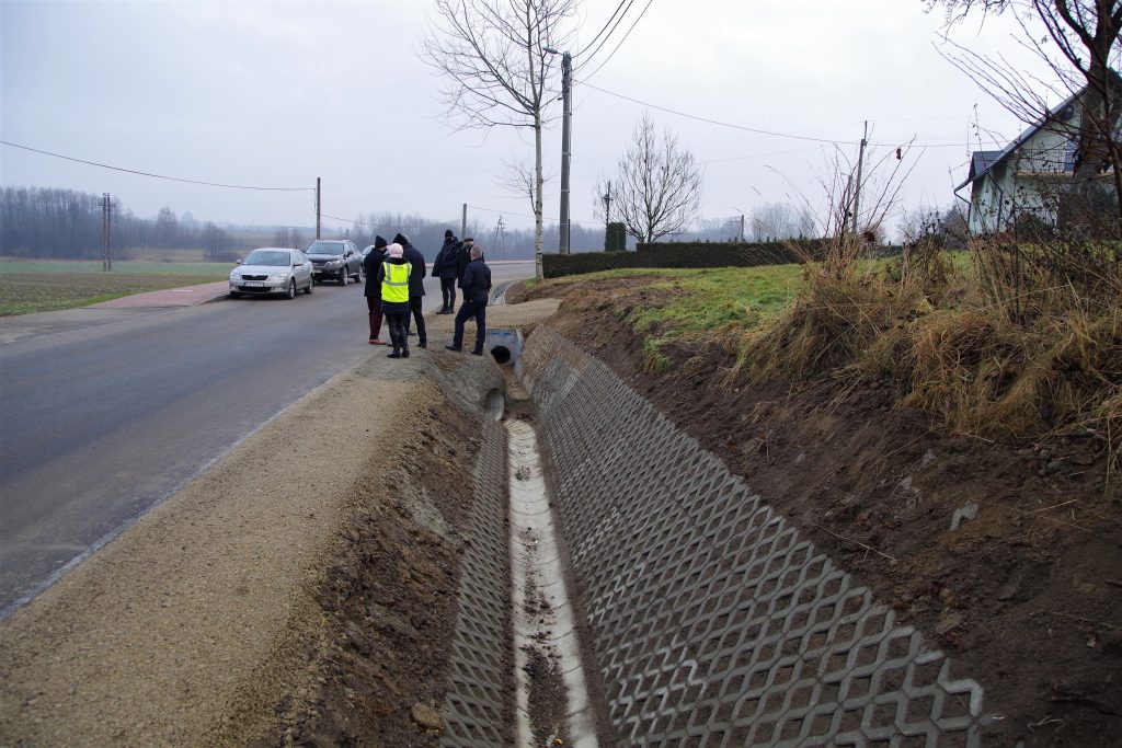 Jeden z rowów przy przebudowanej drodze, wzmocniony betonowymi kratkami, W tle grupa osób.