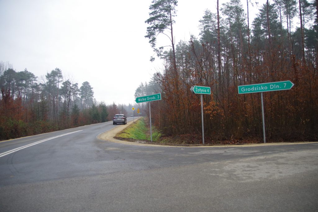 Skrzyżowanie kończące przebudowę drogi. Na poboczu znajdują się trzy zielone znaki wskazujące drogi do poszczególnych miejscowości: Wólki Grodziskiej, Grodziska Dolnego oraz Żołyni.
