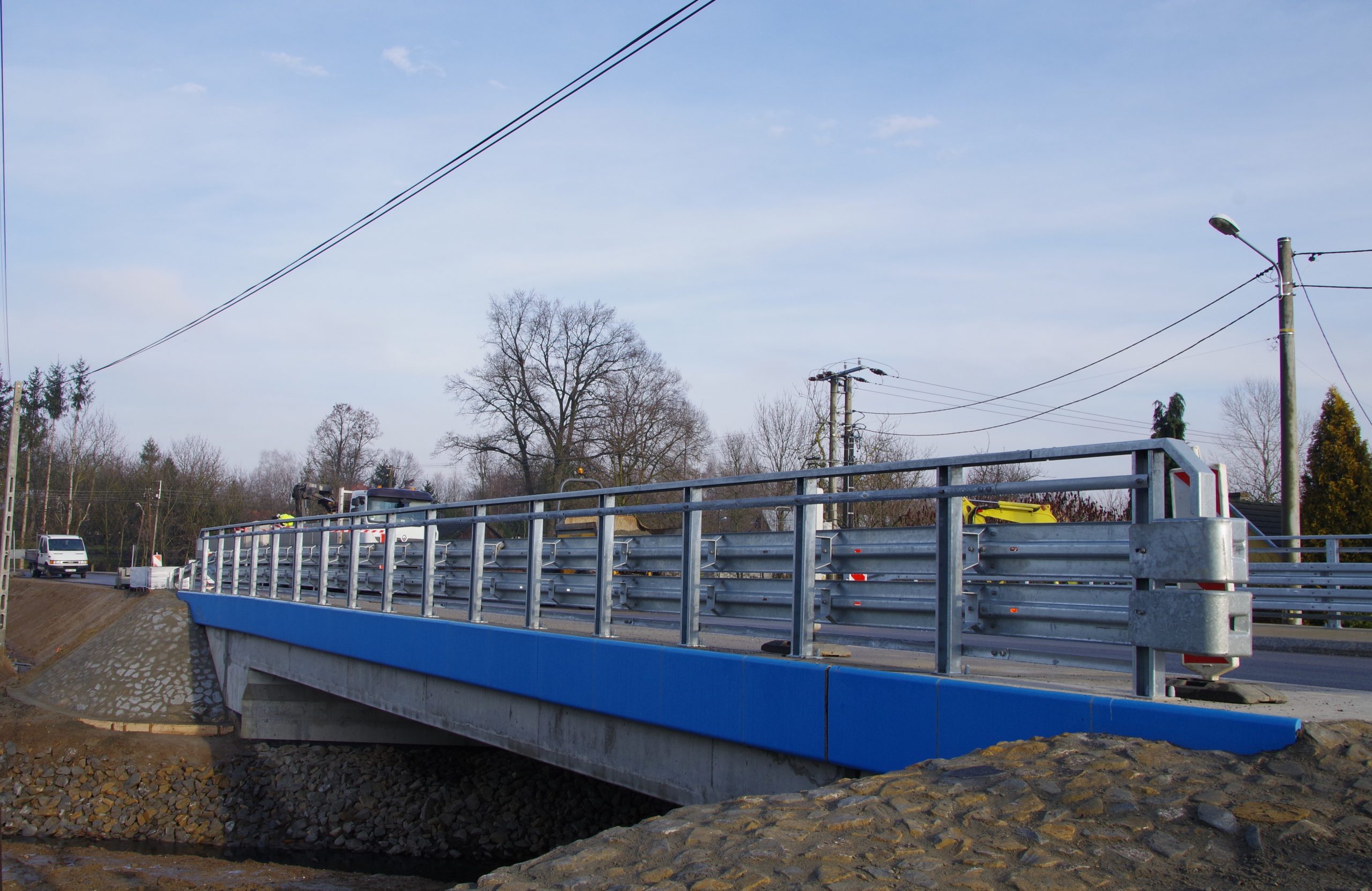 Most w Grodzisku Dolnym po rozbudowie. Metalowe przęsła, boki mostu na wysokości jezdni w kolorze niebieskim, po obu stronach kamieniste zbocza.