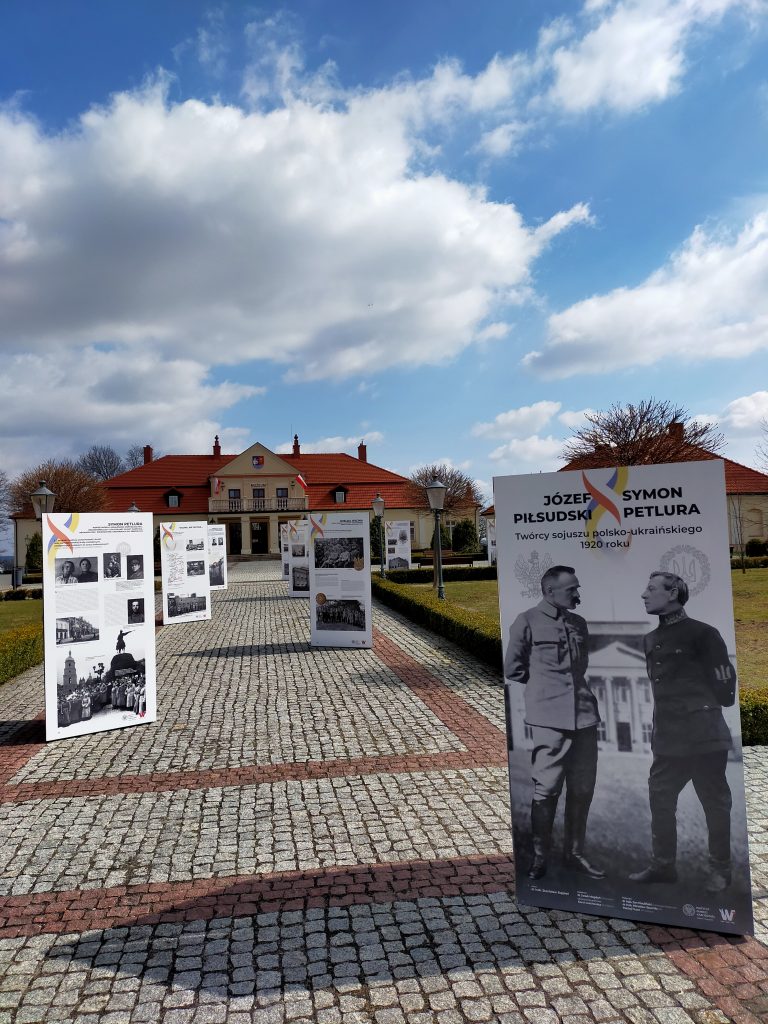 Wzdłuż głównej alejki prowadzącej do muzeum ustawiono biało-czarne plansze z informacjami dotyczącymi postaci Piłsudskiego i Petlura. W tle widoczny budynek muzeum.