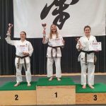 Trzy kobiety stojące na podium ubrana w białe stroje do karate.