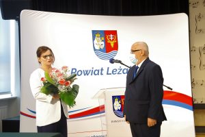 Kobieta w białej marynarce i granatowych spodniach oraz mężczyzna w ciemnoszarym garniturze stoją na tle białego baneru z herbem powiatu i napisem Powiat Leżajski. Kobieta trzyma bukiet kwiatów.