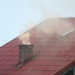 Fragment czerwonego dachu domu jednorodzinnego z widocznym na nim kominem, z którego unosi się gęsty dym.