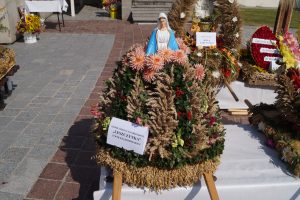 Wieniec dożynkowy wykonany ze zbóż. Kształtem przypomina stożek, na którego szczycie znajduje się gipsowa figurka Matki Boskiej w biało-niebieskich szatach.