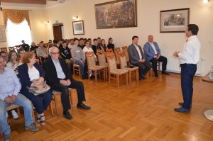 Wnętrze sali konferencyjnej, na pierwszym planie publiczność, w sali panuje żółte światło.