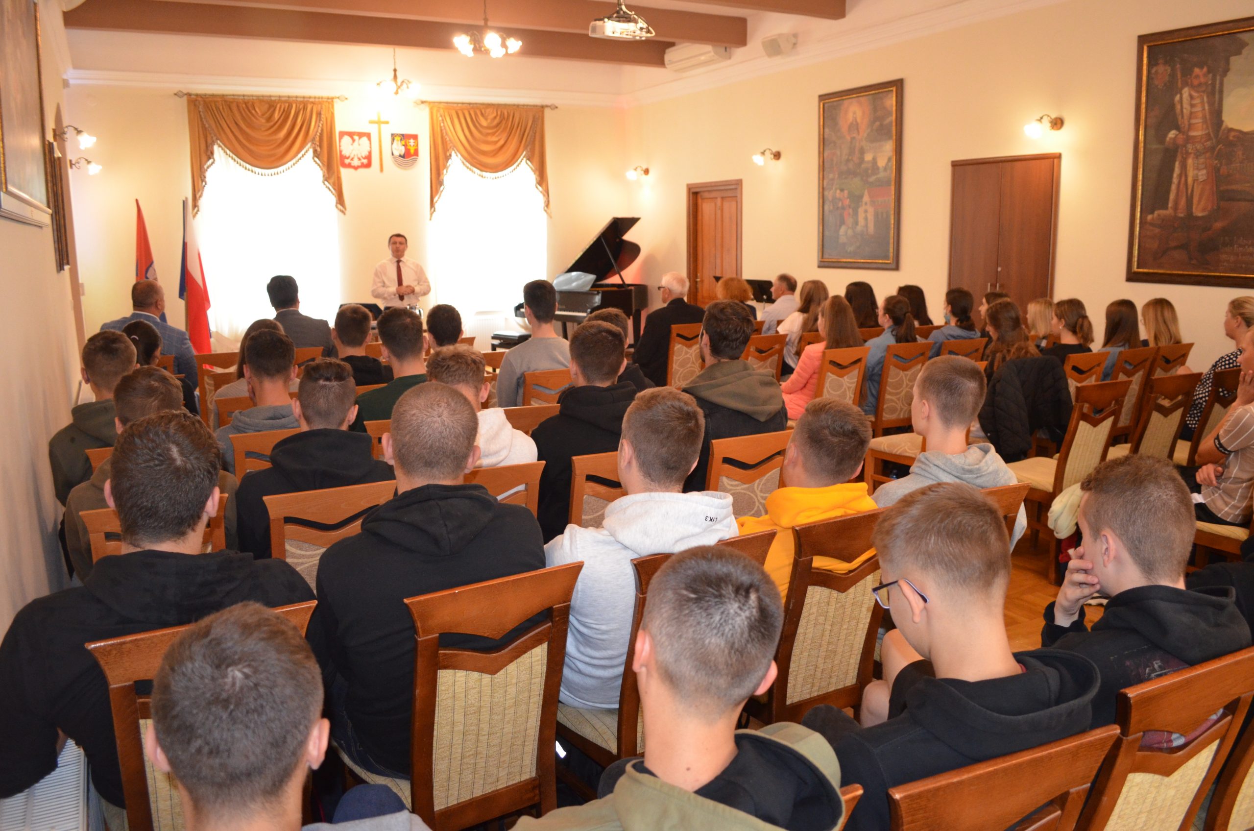 Uczniowie szkół średnich siedzą na sali konferencyjnej muzeum, widok z końca sali, w tle osoba przemawiająca, w sali panuje żółte światło.