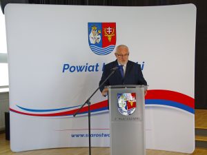 Mężczyzna w czarnym garniturze przemawia przy mównicy na tle białej ścianki, na której znajduje się herb powiatu leżajskiego i napis Powiat Leżajski w barwach czerwono-niebieskich.