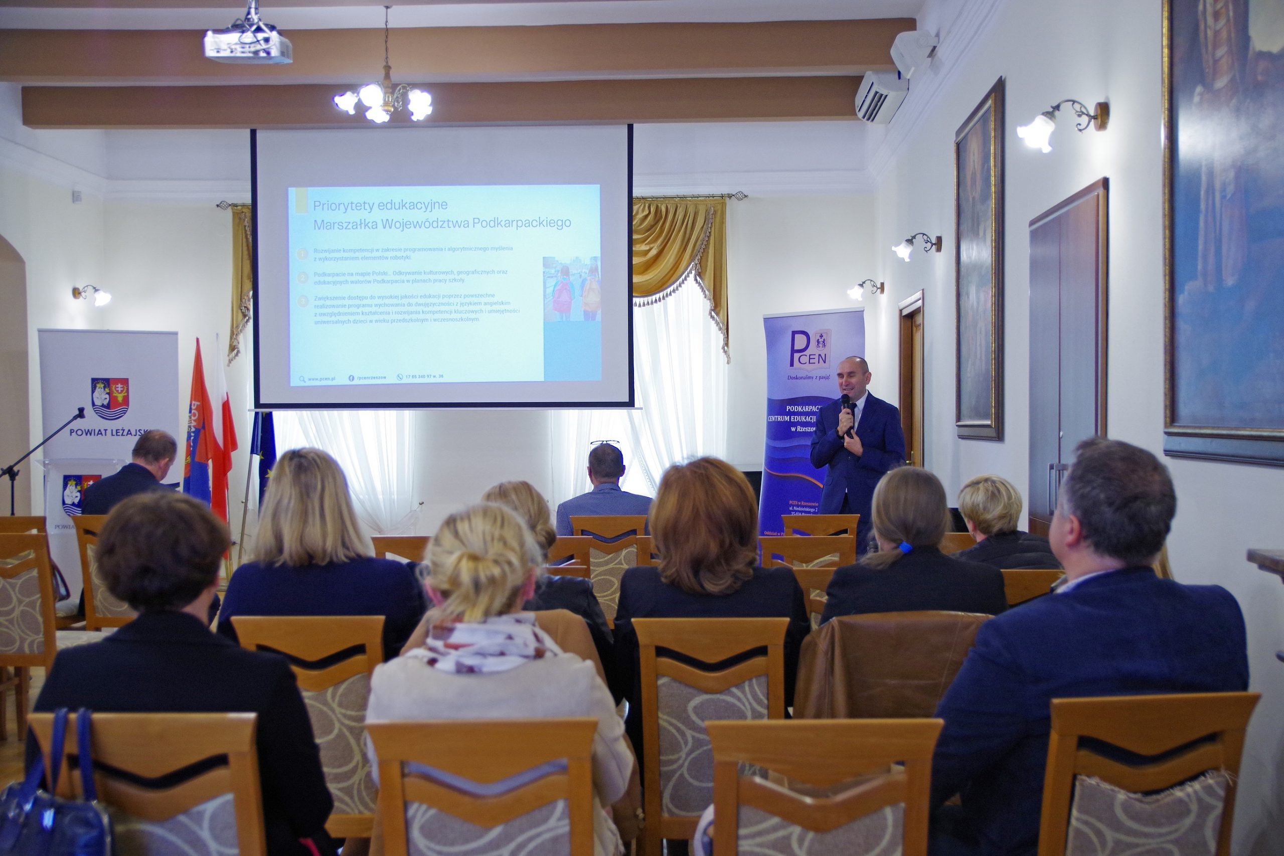 Na pierwszym planie siedzą uczestnicy konferencji zwróceni tyłem do obiektywu, a przodem do przemawiającego mężczyzny. Za mężczyzną wisi ekran projektora, na którym wyświetlana jest prezentacja.