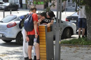 Trzech chłopców pochyla się nad ulotką z questem. W tle białe auto, drzewo i wejście do urzędu miasta.