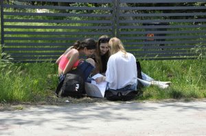 Trzy dziewczyny siedzą na trawie i pochylają się nad ulotką z questem, w tle szare ogrodzenie.