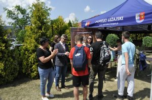 Grupa ludzi stoi, na pierwszym planie kobieta, obok niej mężczyzna, w tle inna kobieta. Na przedzie uczniowie z plecakami zwróceni tyłem do zdjęcia odbierają nagrody.