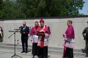 Trzech księży oraz mężczyzna w garniturze stoją przed pomnikiem, jeden z księży przemawia.