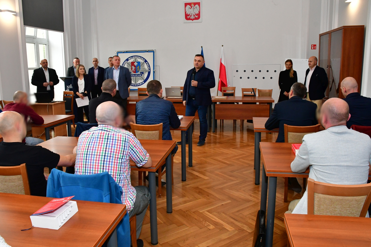 Mężczyzna przemawia do grupy mężczyzn. Po prawej stoi pięciu mężczyzn i jedna kobieta. Po lewej stoi jedna kobieta i jeden mężczyzna.