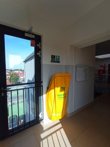 Krzesło ewakuacyjne pokryte żółtym pokrowcem umocowane na ścianie - widok od strony windy.