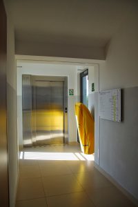 Krzesło ewakuacyjne okryte żółtym pokrowcem umieszczone na ścianie przy wejściu do windy - widok od strony korytarza.