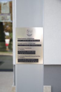 Tabliczka informacyjna umieszczona na filarze przed głównym wejściem do starostwa. Tabliczka zawiera informacje w języku polskim oraz Braille'a.